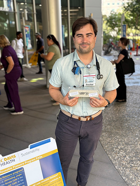 An adult holds up a reusable utensil kit next to a sign that promotes sustainability at UC Davis Health. 
